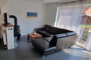 A living room with a sofa, a small table, a heater, and a window with a view of a building. - Wismar cog starboard (Zierow)