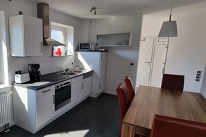 A compact kitchen with white cabinetry, a microwave, a toaster, and a stove. There is a dining table with chairs and a pendant light. - Wismar cog starboard (Zierow)