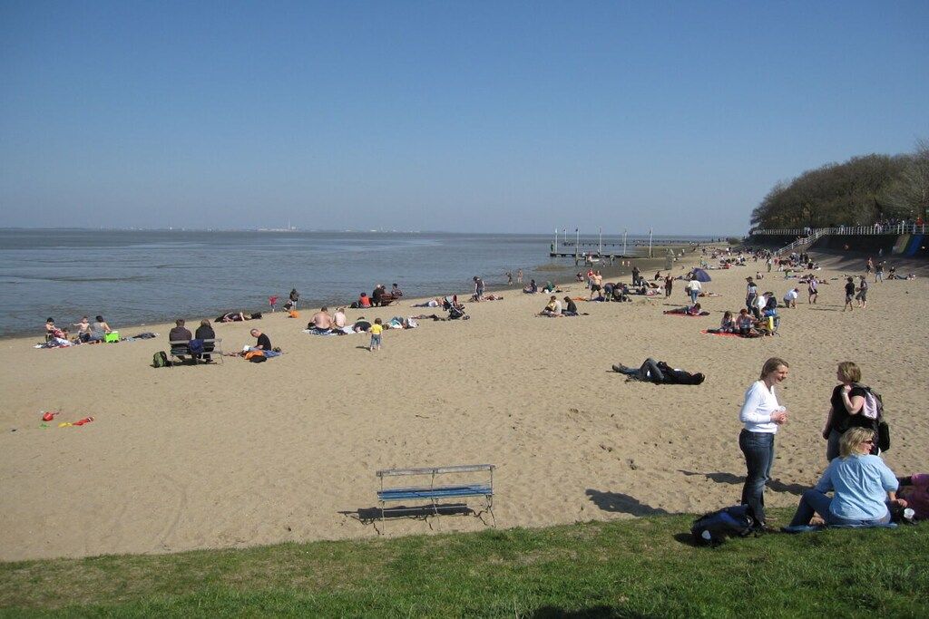 En strand där folk solar och promenerar, ett grönområde med en bänk och en pir i bakgrunden.