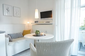 Ein modernes Wohnzimmer mit einem weißen Sofa, einem runden Tisch mit einer Pflanze, einem an der Wand montierten Fernseher und einem großen Fenster mit durchsichtigen Vorhängen.