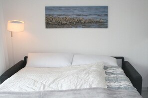 Ein Bett mit weißer Bettwäsche und einer grauen Decke, eine Lampe links sowie ein Bild einer Strandlandschaft an der Wand.