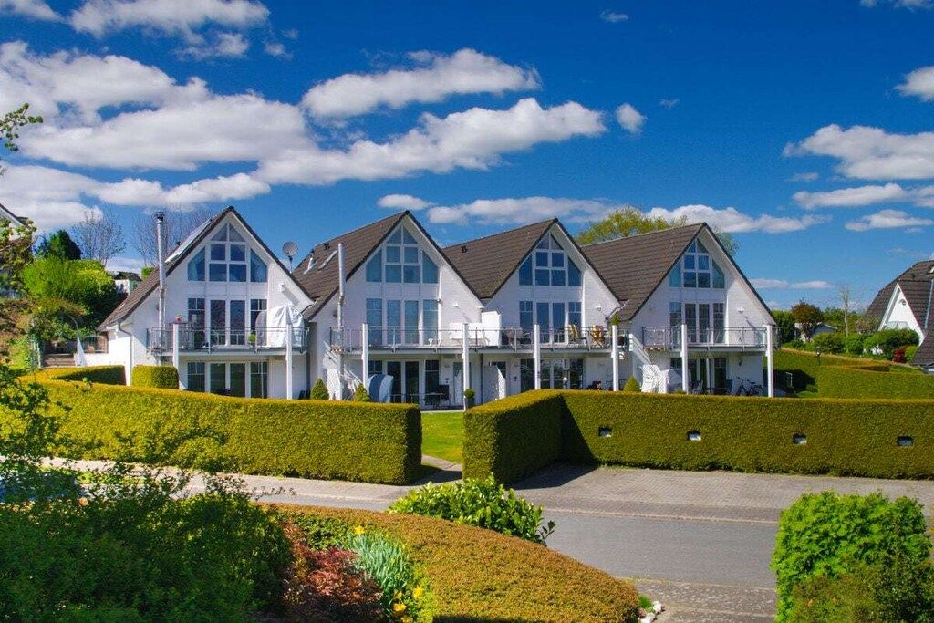 A row of white houses with gabled roofs, surrounded by well-maintained hedges and a clear sky.