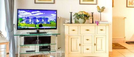 A living room with a television on a glass stand, a white cabinet with drawers, and framed pictures on the wall.