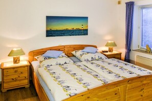 A double bed with a nautical-themed duvet, two bedside lamps, a nightstand with a drawer, a framed picture of a lighthouse on the wall, and a window with curtains. - Holiday apartment with 1 bedroom (Lindaunis)