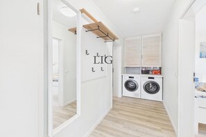 A compact laundry room with a washing machine, dryer, and a wooden shelf. - Holiday apartment Dünengras (Langeoog)