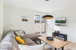 A modern living room with a sofa, a coffee table, a television mounted on the wall, and a pendant light. - Holiday apartment Dünengras (Langeoog)