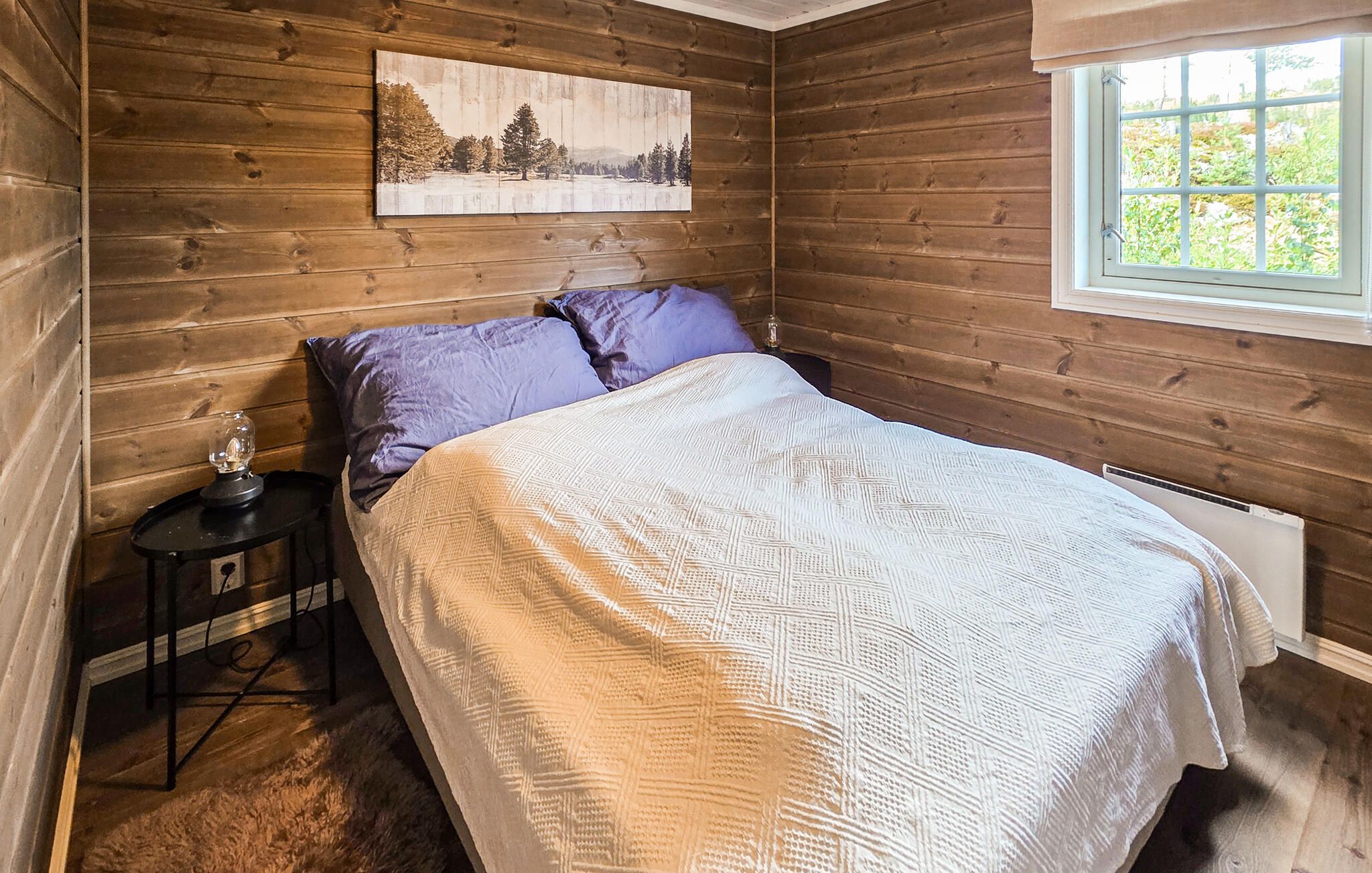 Een slaapkamer met een houten wand, een bed met een wit geborduurde spreideken, twee kussens, een nachtkastje met een lamp en een raam met uitzicht op groen.