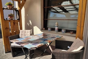 Ein Außenbereich mit Sitzgelegenheit, einem Tisch mit Patchwork-Tischdecke, Korbstühlen und Kübelpflanzen auf einem Balkon.