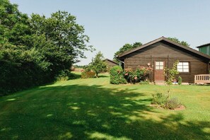 Eine Holzhütte mit geneigtem Dach, umgeben von einer gepflegten Rasenfläche und dichtem Strauchwerk.