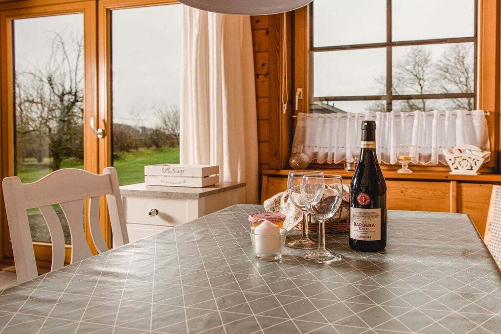 A table with a bottle of wine, glasses, and a small container, set against a window with a view of trees.