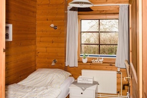 A wooden cabin bedroom with a bed, a bedside table with a lamp, a window with curtains, and a radiator. - Struve 1 on the Kastanienhof (Quern)
