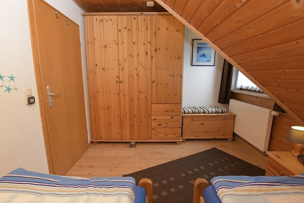 A bedroom with a wooden wardrobe, a bedside table, a bed with striped bedding, and a window with a view.