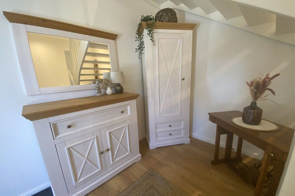 Habitación de esquina con una consola de madera, un armario blanco con cajones, un espejo y una mesita con un florero.