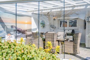 A glass-enclosed patio with wicker furniture, a television, and a view of the sea. - House Sansibar Niendorf (Timmendorfer Strand)