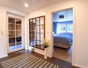 A hallway with a patterned rug, a wooden-framed mirror, and a door leading to a bedroom with a bed and window. - Holiday Home 'Holidate-Inn' with Private Terrace, Private Garden and Wi-Fi (Stadum)