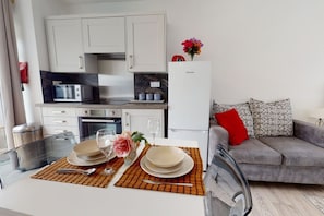 A modern kitchen with white cabinets, a microwave, and a refrigerator. There is a dining table set with plates, glasses, and a flower arrangement. - Apartment 101 - Symons House (Medway)