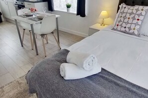 A hotel room with a bed, a bedside table with a lamp, a small table with a chair, and a window with curtains. - Apartment 203 - Symons House (Medway)