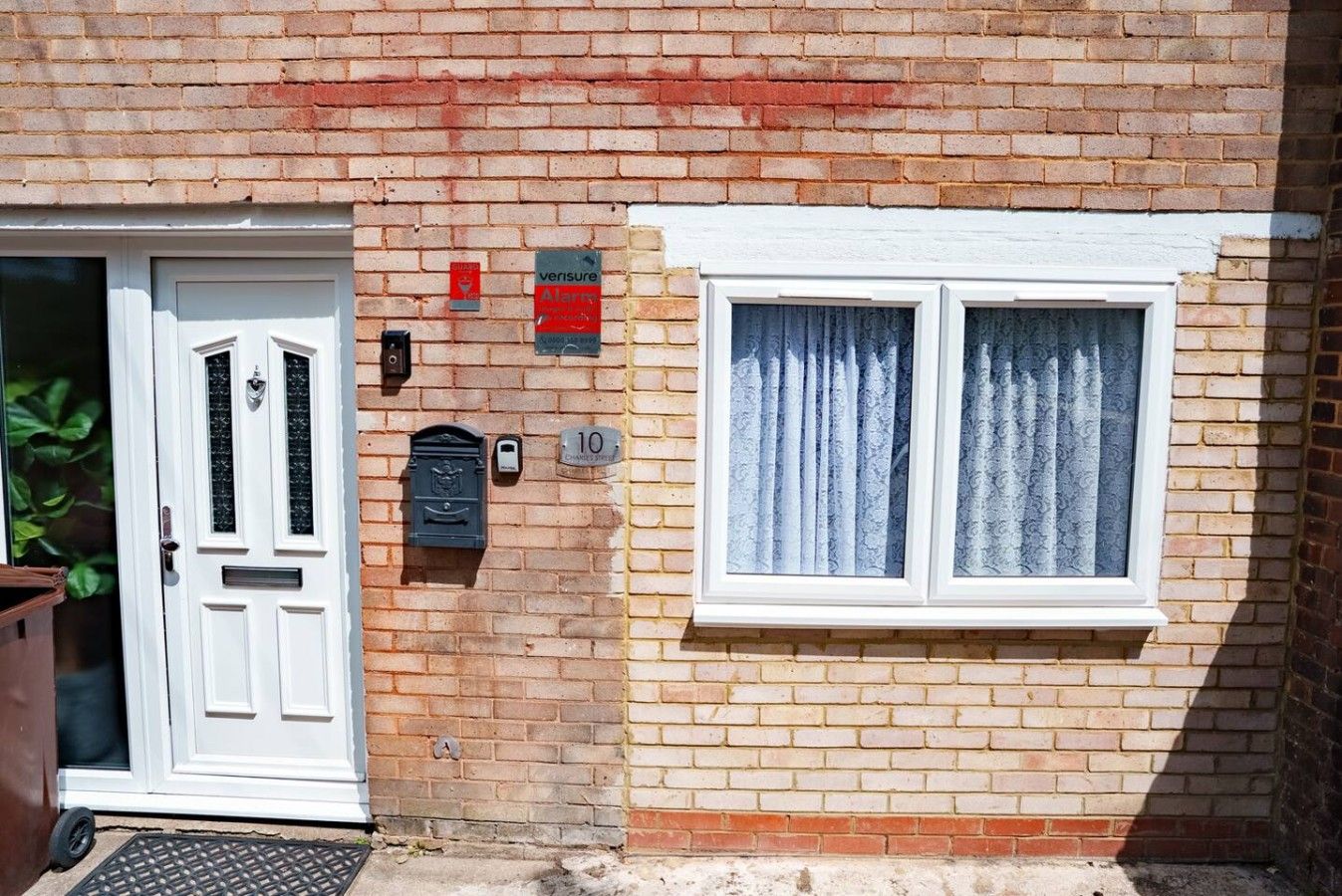 A brick house with a white door, a mailbox, a number plate, and a window with curtains.