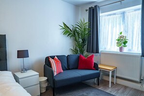 Ein modernes Schlafzimmer mit Bett, Sofa, Nachttisch, Lampe, Heizkörper, einem Fenster mit Vorhängen und einer Topfpflanze.