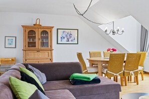 Ein modernes Wohnzimmer mit einer Eckcouch, einem Esstisch mit Korbstühlen, einem Holzschrank und einer Kronleuchter.