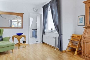Ein Zimmer mit einem grünen Sofa, einem Holzstuhl, einem kleinen Tisch mit einer blauen Schale, einem Spiegel, einem Fenster mit Vorhängen und einer Heizung.