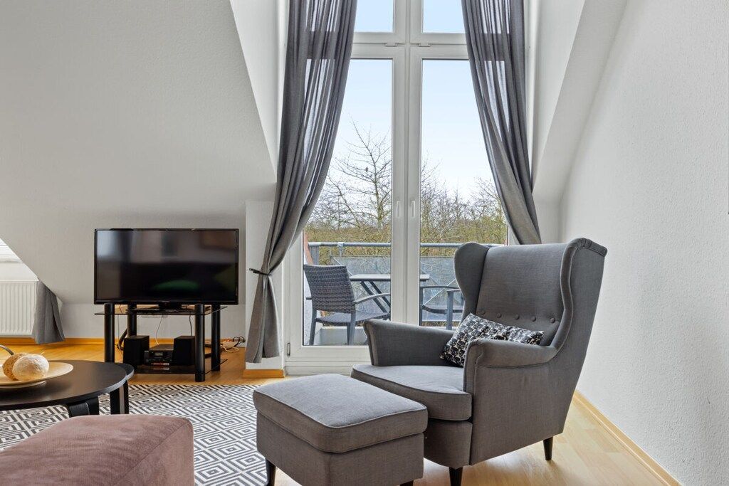 A modern living room with a flat-screen TV, grey armchair, ottoman, and a patterned rug.