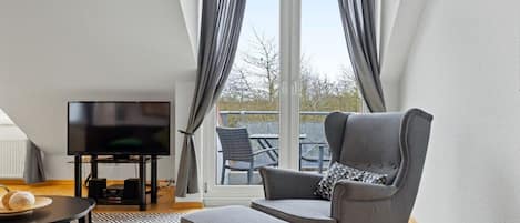 A modern living room with a flat-screen TV, grey armchair, ottoman, and a patterned rug.