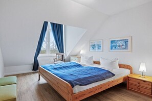 Ein Schlafzimmer mit einem hölzernen Bett, einem Nachttisch, einem Sessel, einem Fenster mit Vorhängen und zwei gerahmten Bildern an der Wand.