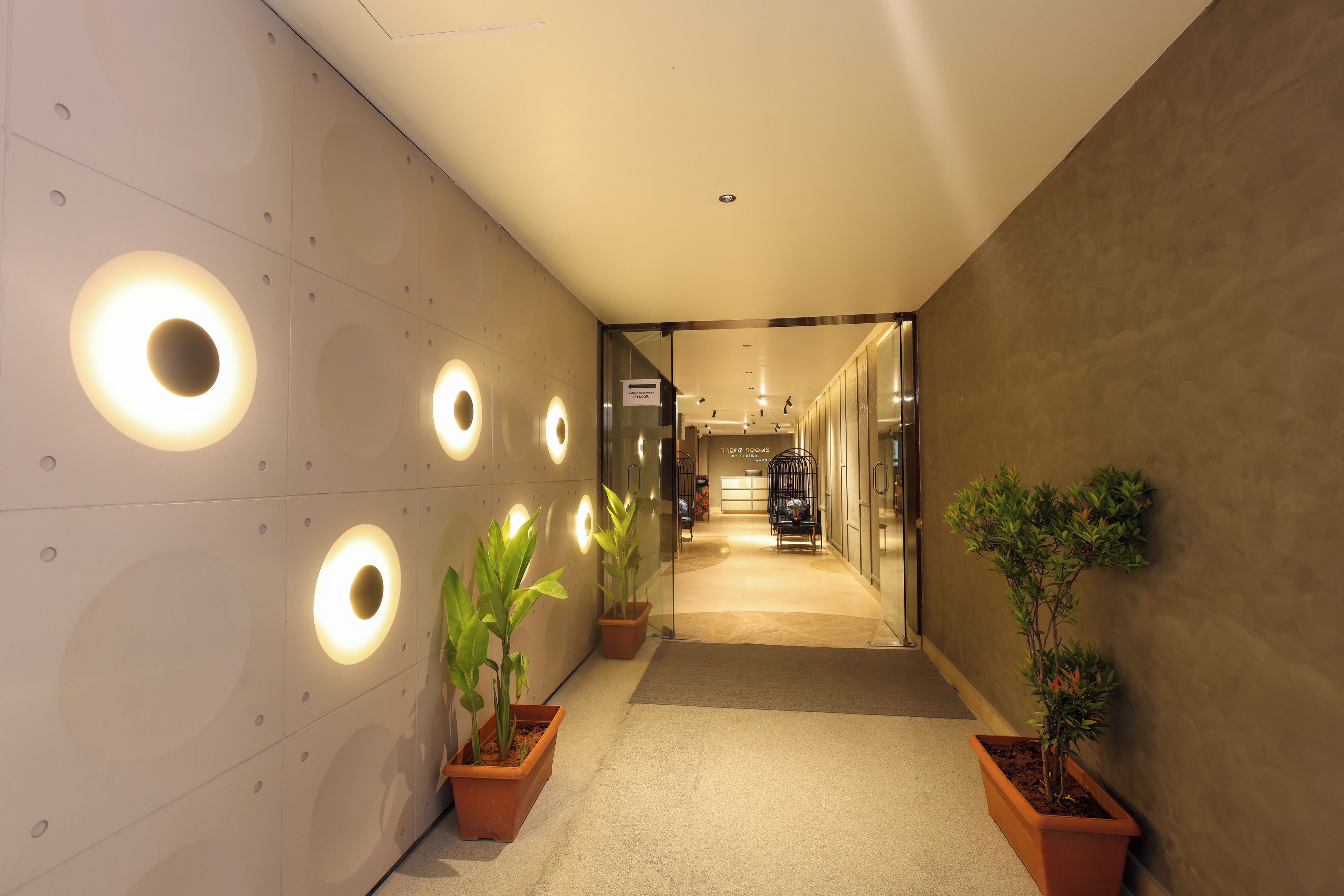 Un couloir orné de luminaires circulaires fixés au mur, de plantes en pot et d’un panneau indiquant la direction à suivre pour accéder au troisième étage.