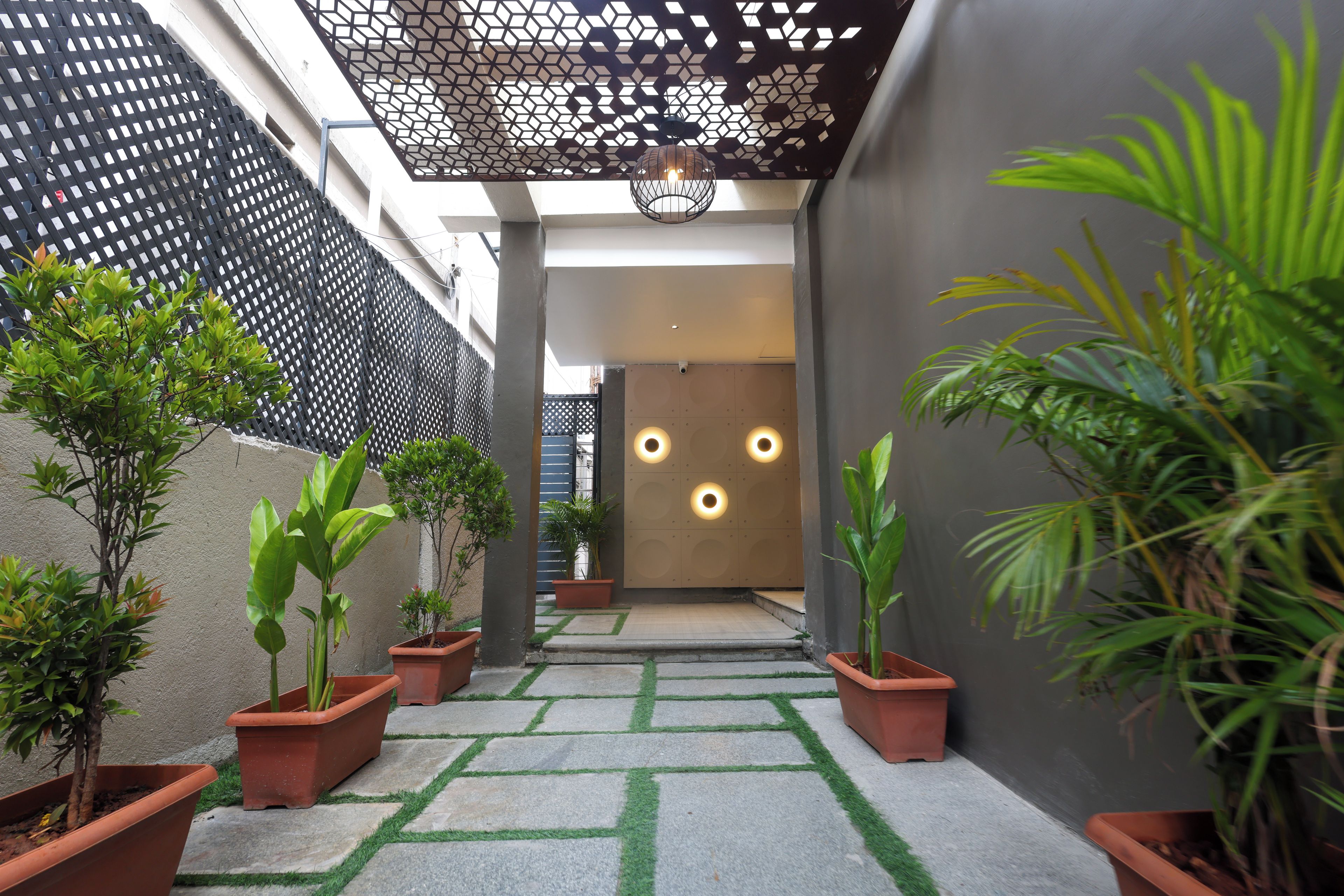 Un sendero estrecho bordeado de plantas en macetas que conduce a la entrada de un edificio con techo de diseño geométrico e iluminación empotrada.