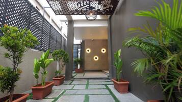 Un étroit sentier bordé de plantes en pot mène à l’entrée d’un bâtiment, dont le plafond présente un motif géométrique et qui est doté d’un éclairage encastré.