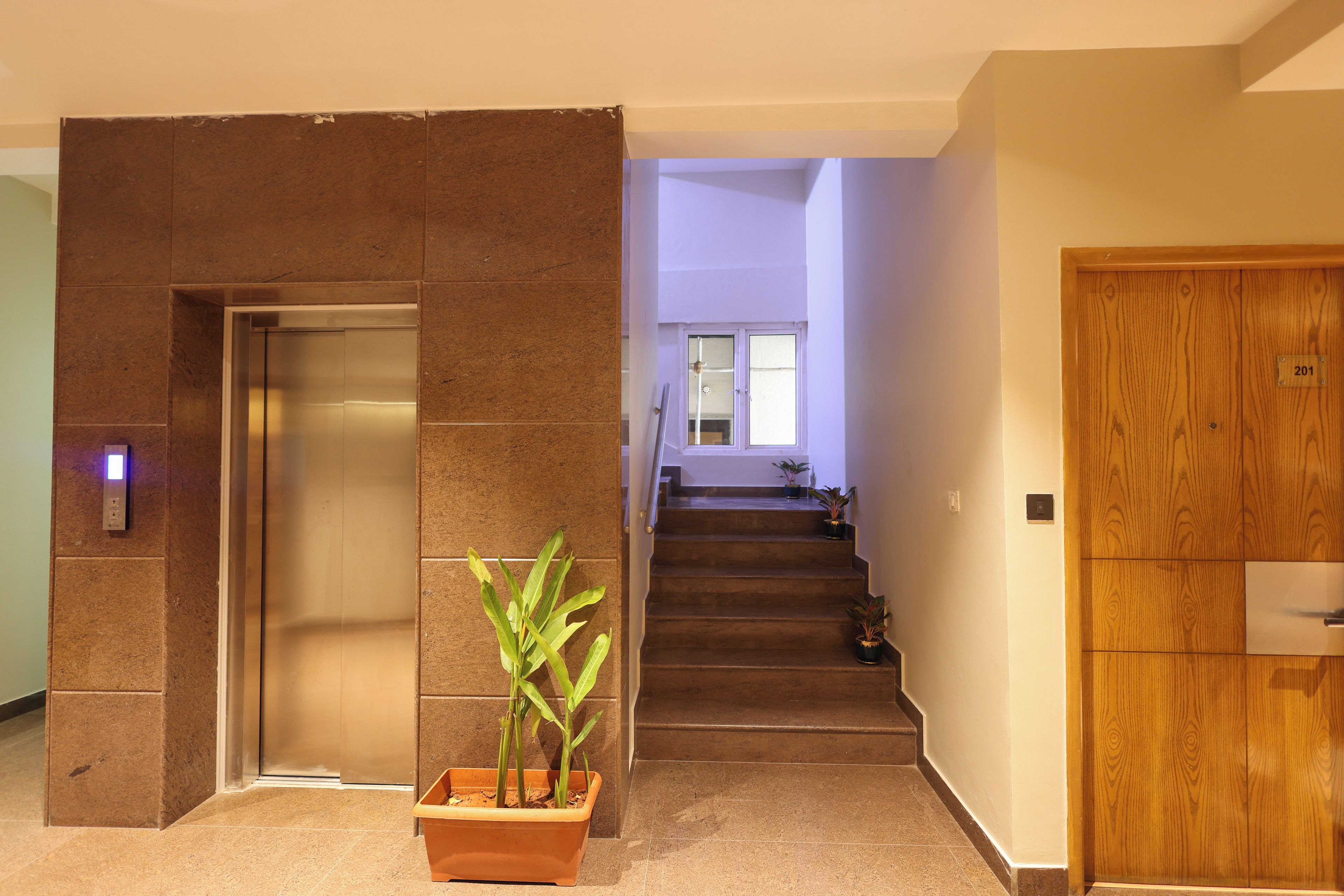 Un intérieur moderne avec un ascenseur fixé au mur, un escalier menant à l’étage supérieur et une porte en bois avec une plaque numérotée.