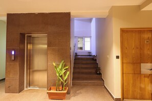 Un interior moderno con un ascensor de pared, una escalera que conduce a un piso superior y una puerta de madera con una placa de número.