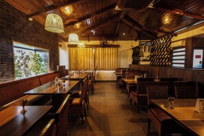 Interior de restaurante con mesas y sillas de madera, lámparas colgantes y vista a zonas verdes a través de una ventana.