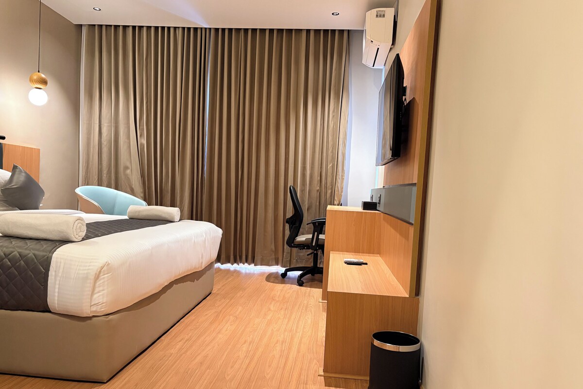 Habitación de hotel moderna con cama, escritorio, silla, televisor y cubo de basura.