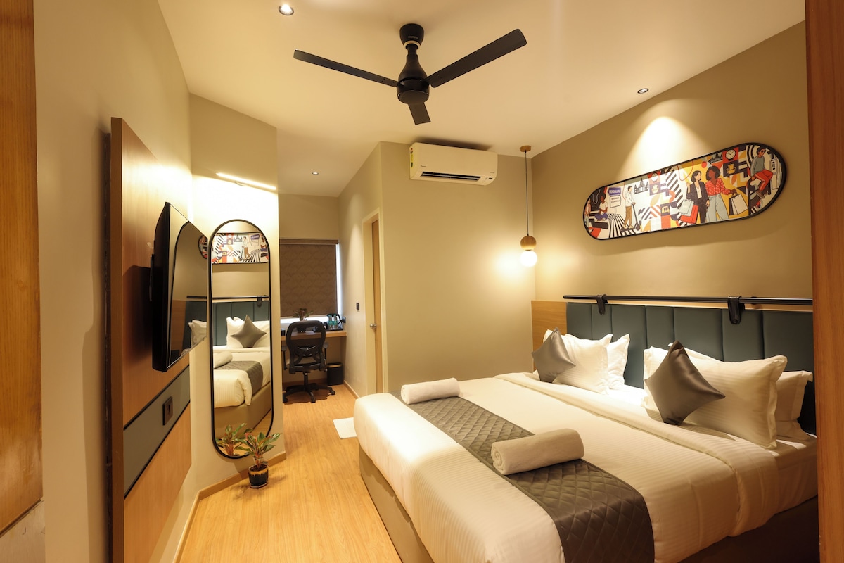 Habitación de hotel con cama, escritorio, silla, ventilador de techo, unidad de aire acondicionado, televisor montado en la pared y un adorno decorativo.