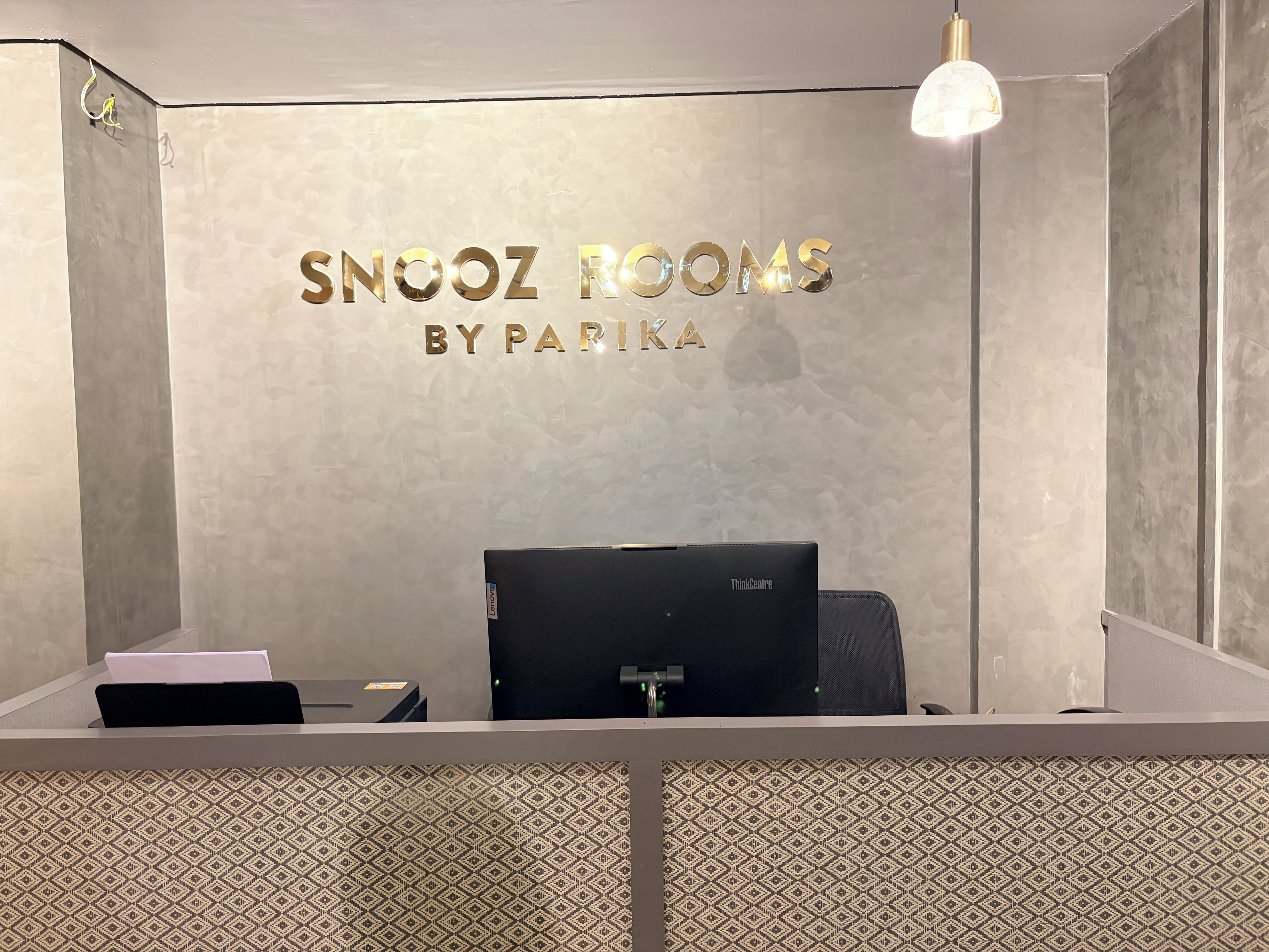 A reception desk with a laptop, a pink folder, and a black chair. The wall behind has a metallic sign that reads 'SNOOZ ROOMS BY PAPIKA'.