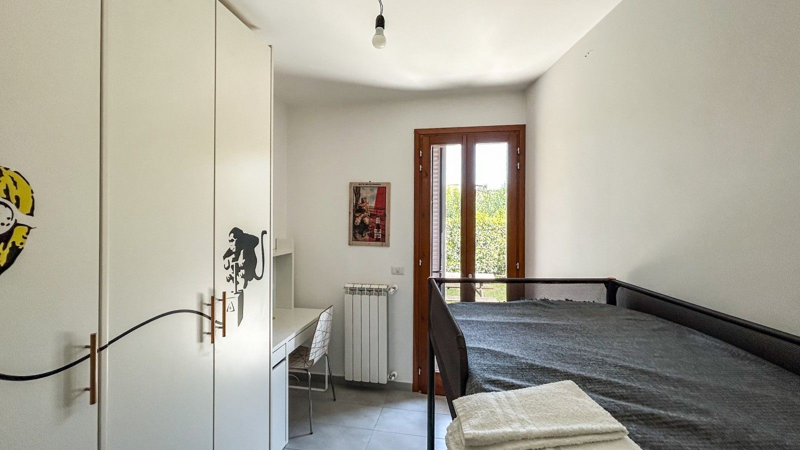 A bedroom with a bed, a desk, a chair, a radiator, and a door leading to a balcony.