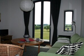 A room with a green sofa, a round wooden table with a teapot and cups, a television on a wooden stand, and a view of a green landscape through a large window. - Holiday apartment with 1 bedroom (Büschow)