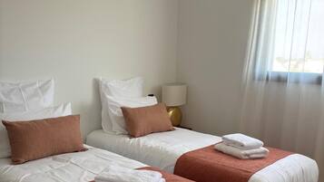 A hotel room with two beds, white bedding, brown throw pillows, a woven light fixture, a bedside lamp, and a window with sheer curtains.