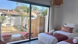 Habitación de hotel con un amplio ventanal corredero que da a un área exterior, una cama con ropa de cama blanca y rosa, y una pequeña mesa redonda con sillas.