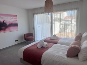 A hotel room with two beds, a chair, a painting, and a large window.