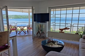 A room with a large window overlooking a body of water, a flat-screen TV mounted on the wall, and a round coffee table with various items on it. - Holiday apartment in Harrislee with Wi-Fi (Harrislee)
