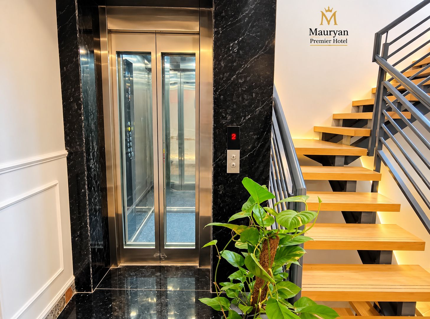 En moderne elevator med digitalt display, en trappe med trindrøjer og metalgelændere, og en potteplante i forgrunden.