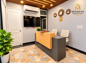Zona de recepción de un hotel con mostrador de recepción, asientos, un reloj en la pared y el logotipo de MAURYAN PREMIER HOTEL.