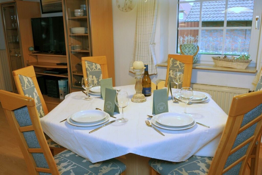 Une table à manger dressée pour quatre personnes, avec des chaises, un téléviseur en arrière-plan et une fenêtre donnant sur une maison.