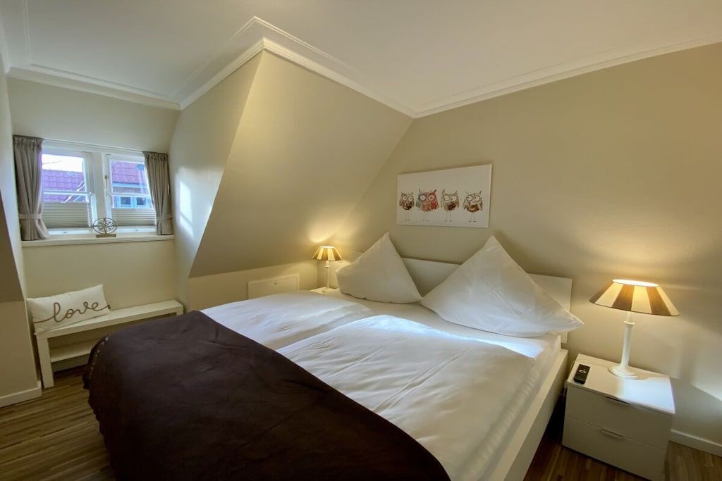 Une chambre à coucher avec un lit, des lampes de chevet, une fenêtre avec des rideaux, une œuvre d’art accrochée au mur et une table de chevet avec une lampe.