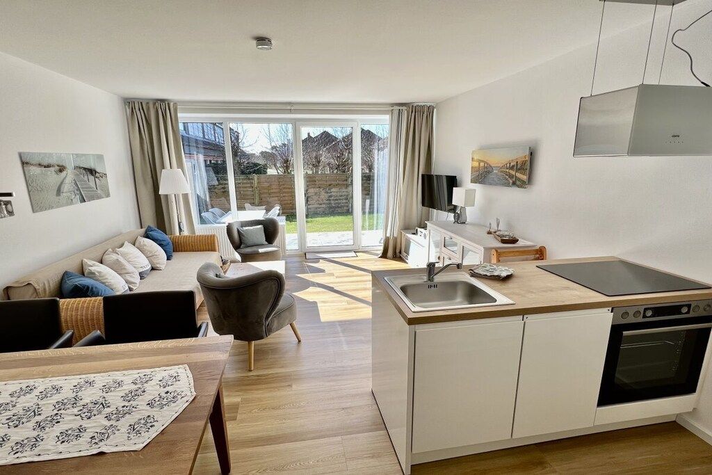 A modern kitchen with a built-in oven, a dining area with a table and chairs, a sofa with cushions, and a view through a glass door to an outdoor area.