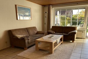 A living room with two brown sofas, a wooden coffee table, and a large window with a view of greenery. - Holiday home with 2 bedrooms (Norddeich)