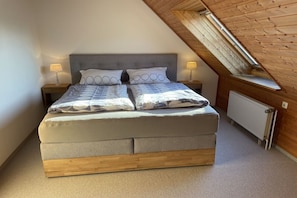 A bedroom with a sloped ceiling, a bed with a grey headboard, two bedside lamps, and a radiator. - Holiday home with 2 bedrooms (Norddeich)
